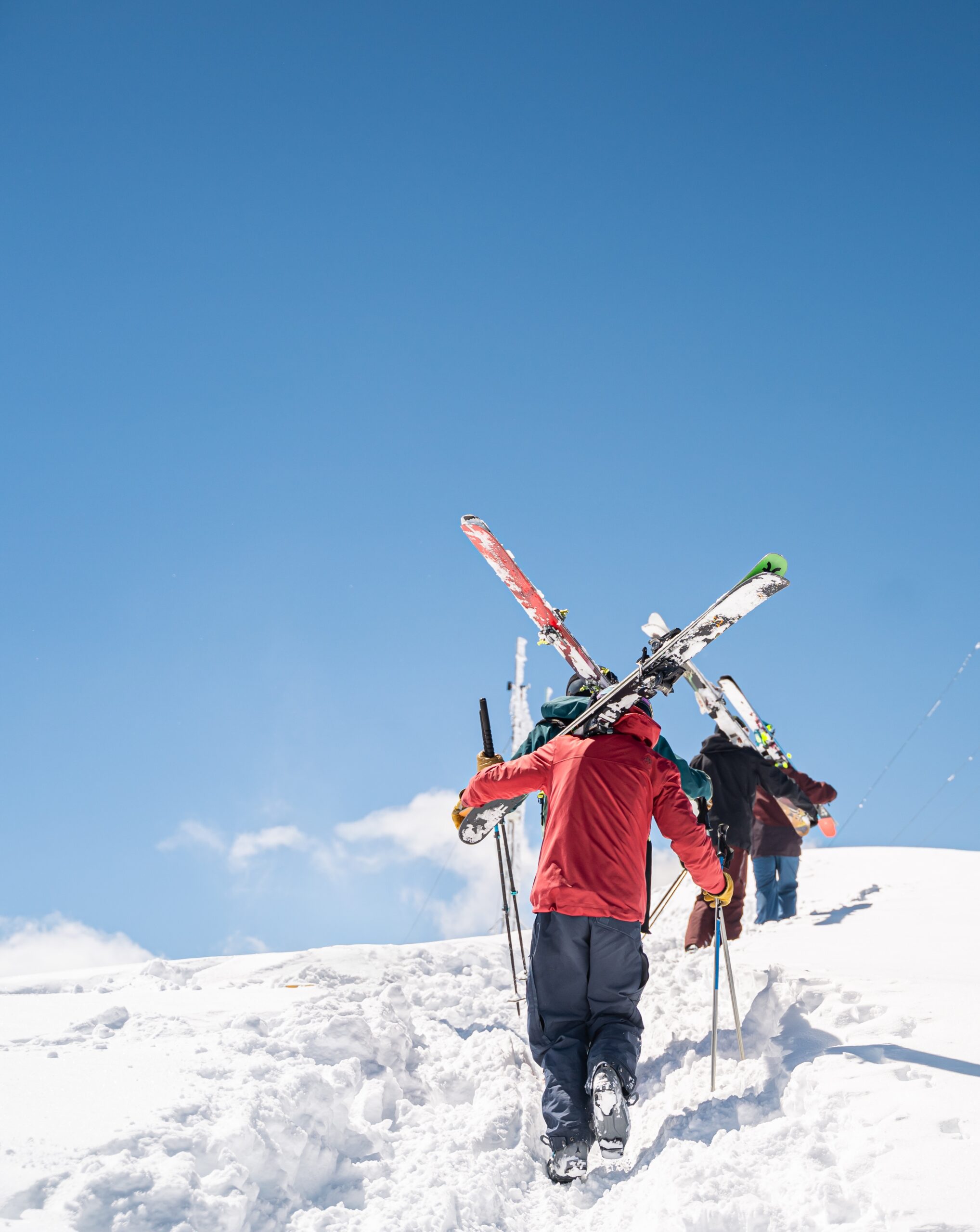 Backcountry skiers hike up a snowy mountain ridge carrying skis under a bright blue sky.