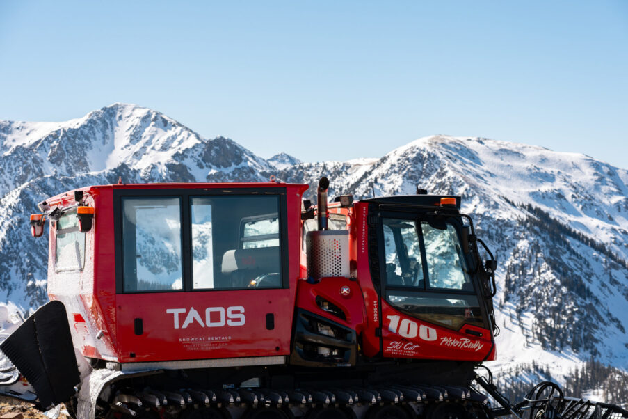 A red snowcat parked on a snowy mountain ridge under a clear blue sky.